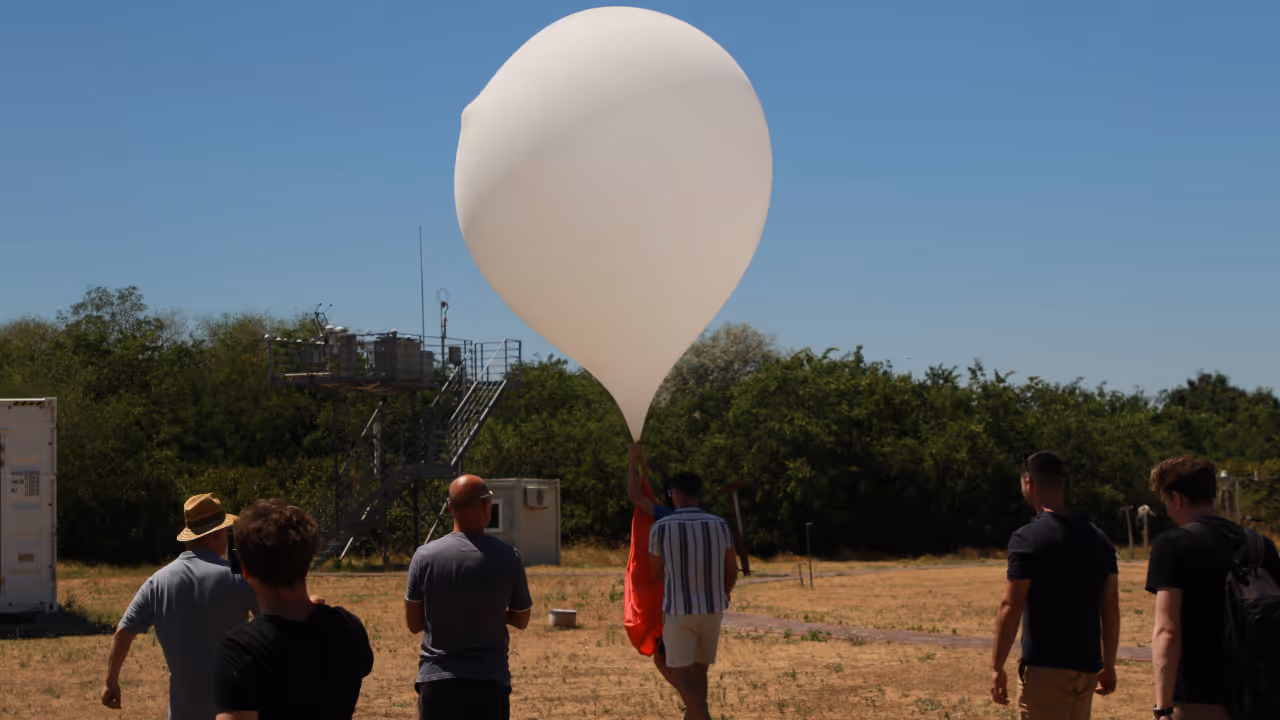Several people walking outdoors on dry grass near a large white weather balloon under a clear blue sky.