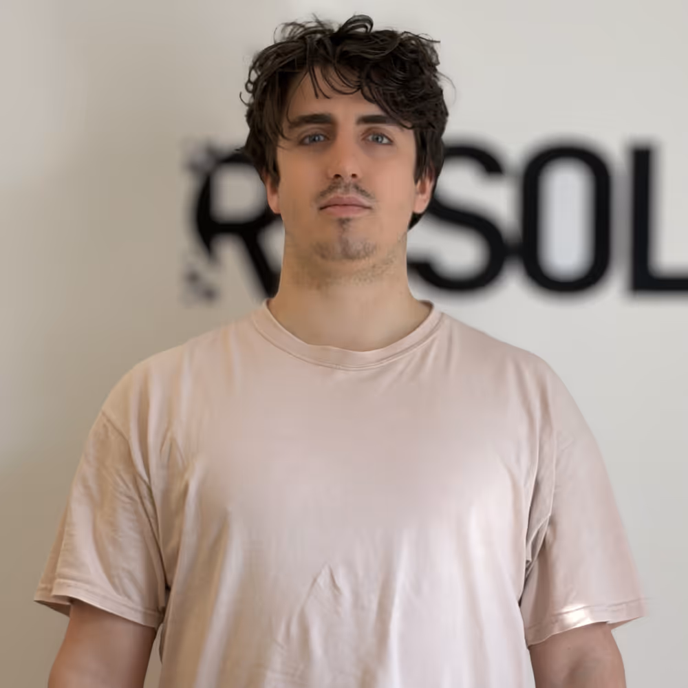 Man with curly dark hair wearing a light pink t-shirt standing in front of a blurred background with text.