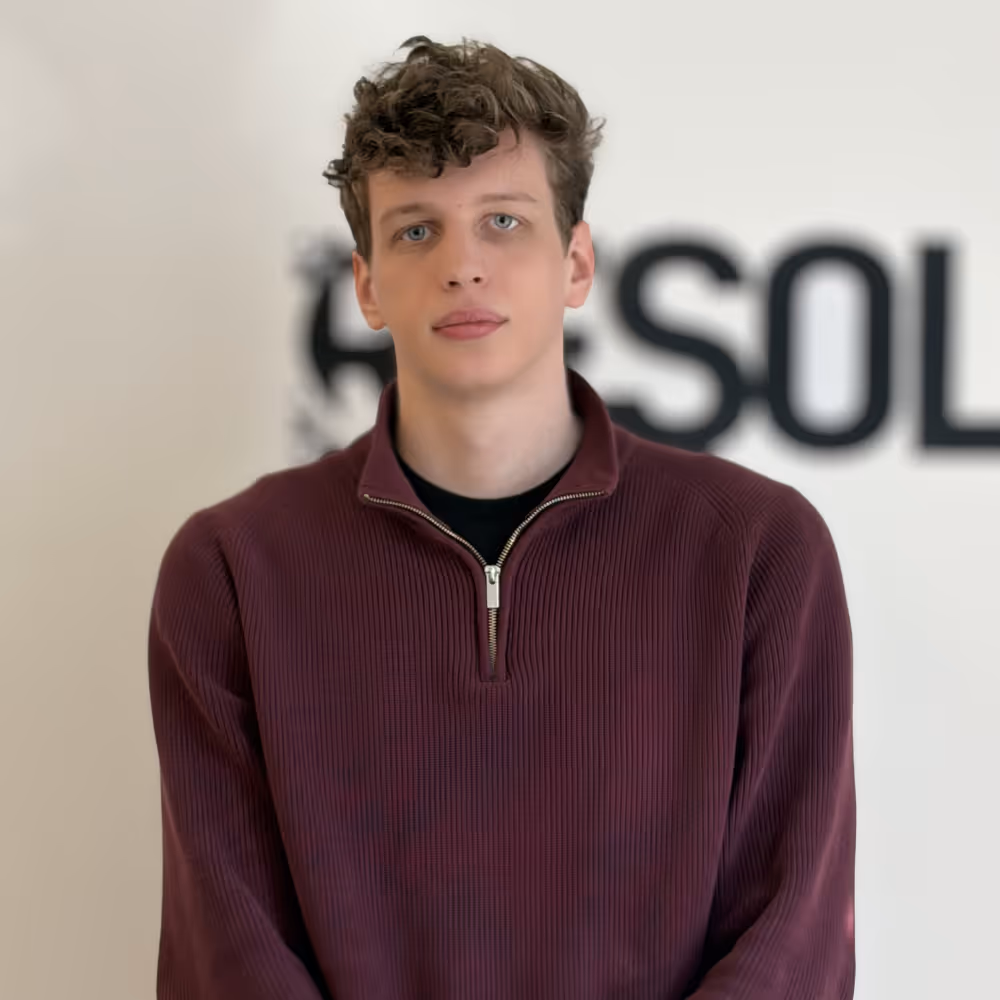Young man with curly hair wearing a maroon zip-up sweater standing against a white wall with blurred black text.