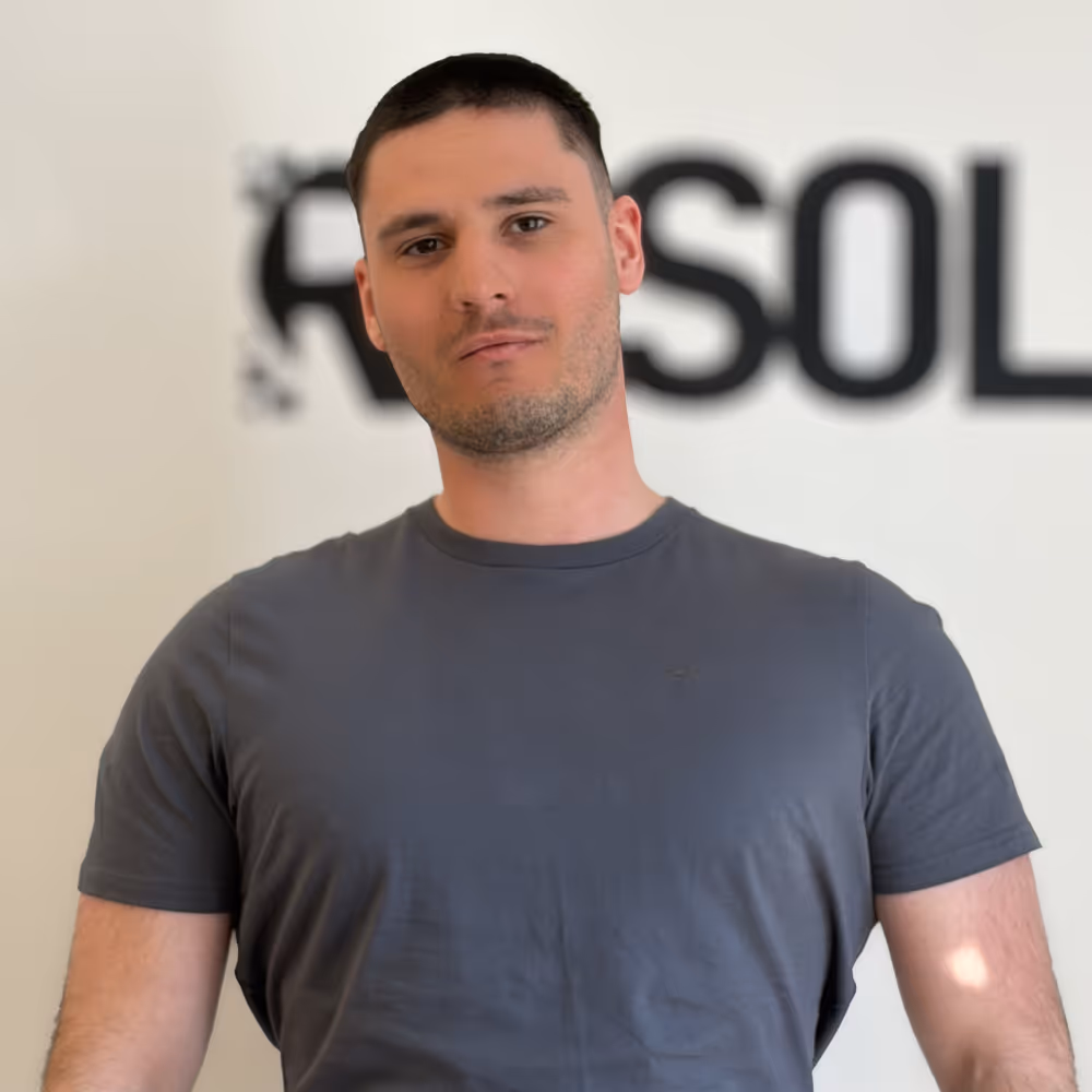 Man with short dark hair wearing a dark gray t-shirt, standing in front of a blurred white wall with partial black text.