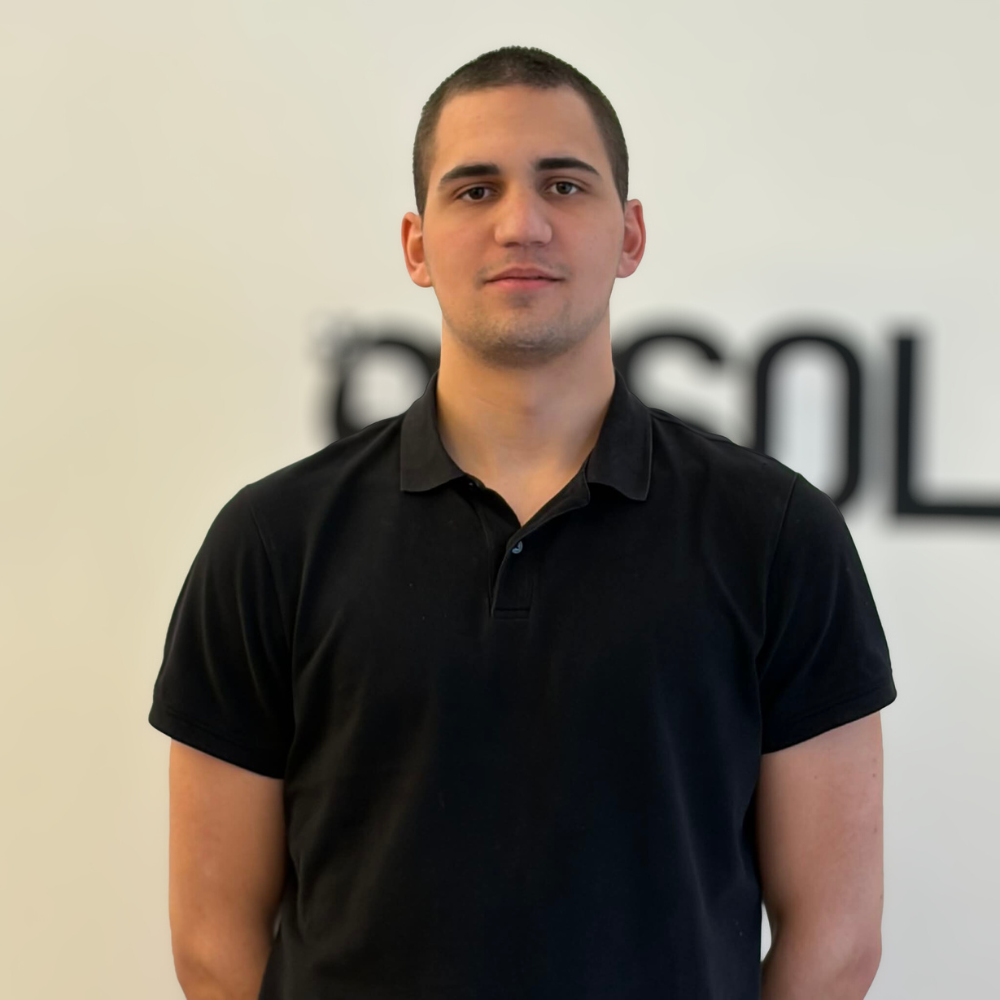 Young man with short dark hair wearing a black polo shirt standing in front of a light wall with blurred text.